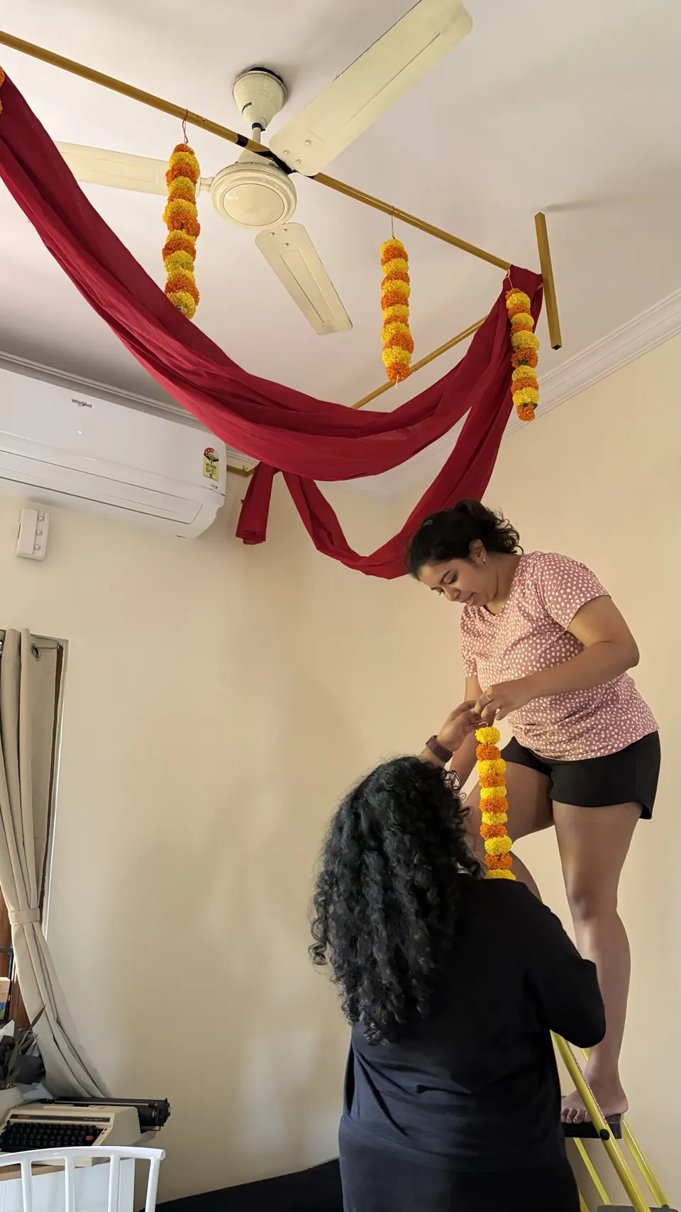 Two women on a ladder hang marigold garlands and maroon drapes from the ceiling before the event
