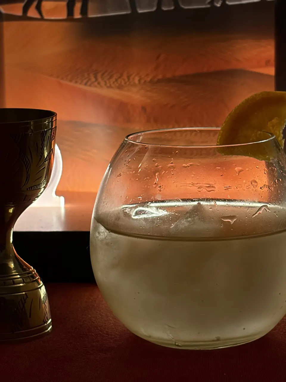 A gin and tonic with an orange wheel in a balloon glass, glowing in front of a Jaisalmer gin desert display beside a brass jigger