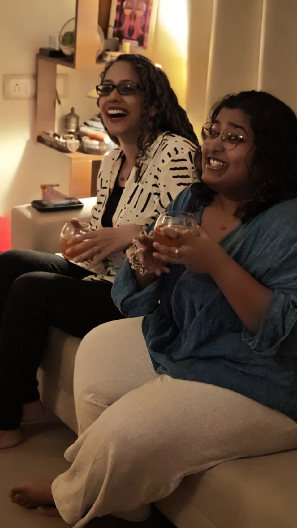 Two guests laughing on a sofa holding rose-petal whisky cocktails in warm lamplight