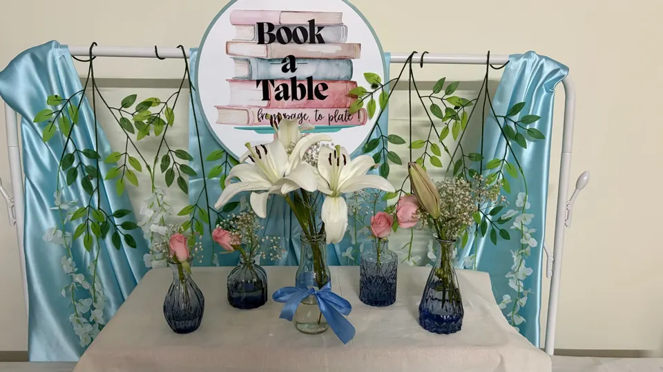 Book a Table sign above white lilies and pink roses in blue glass vases, framed by powder-blue satin drapes and vines