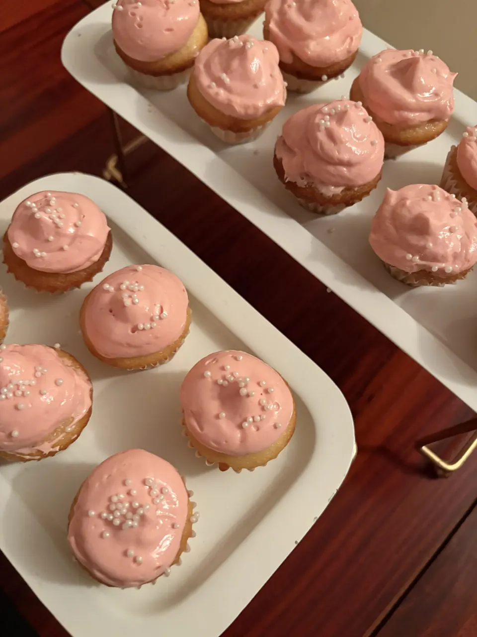 Pink-frosted mini cupcakes topped with white sugar pearls on white trays over a teak table