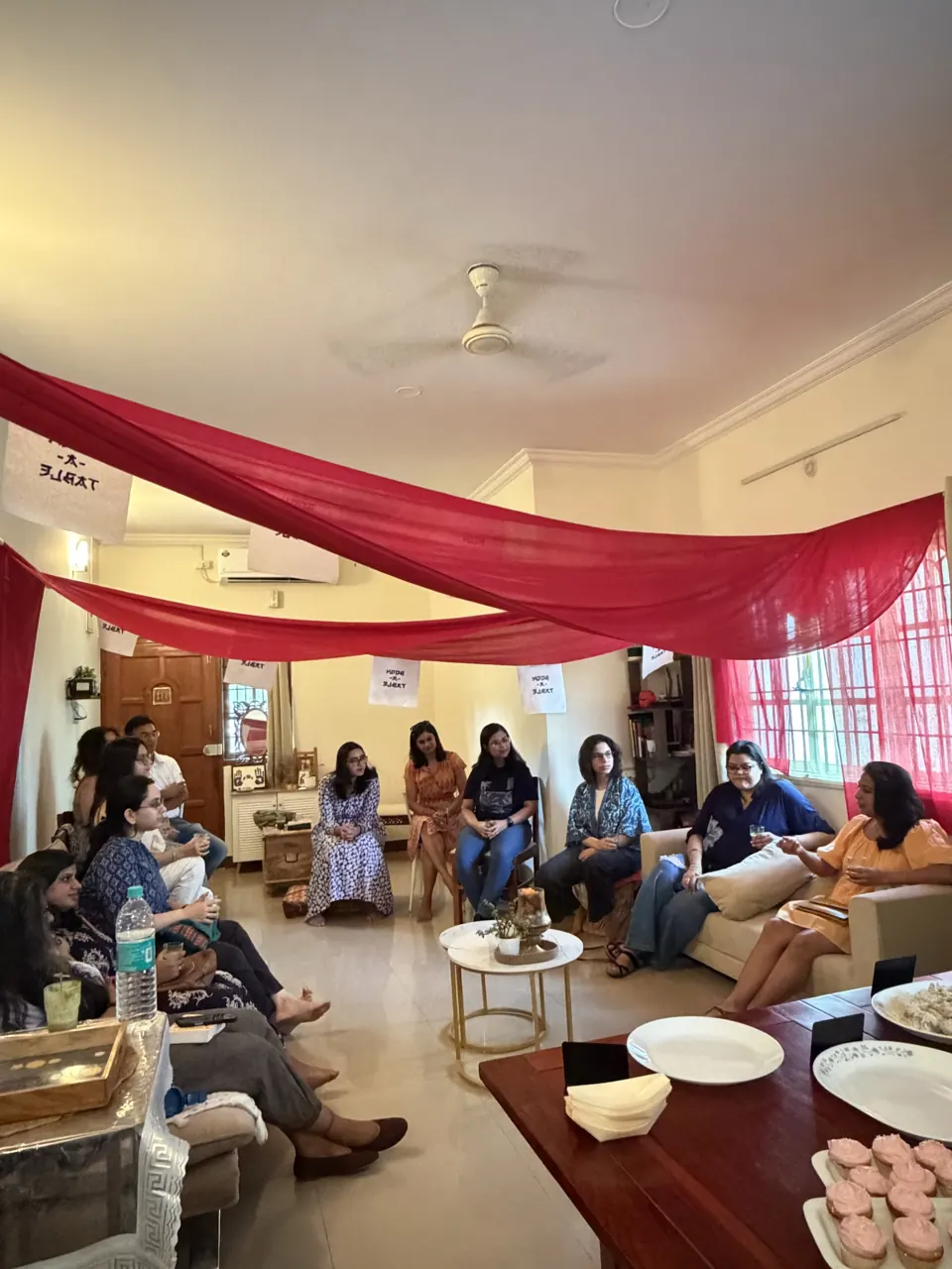 Afternoon light on guests gathered around a living room draped in crimson fabric, plates on the table