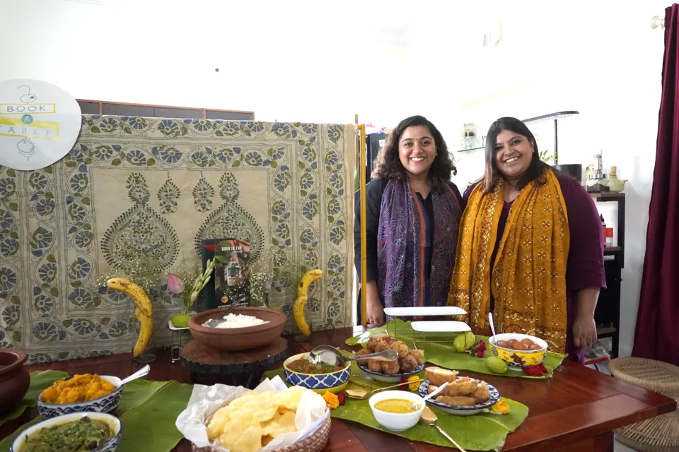 Shachi and Kriti smile behind a banana-leaf Bengali buffet with kantha backdrop and Book A Table sign