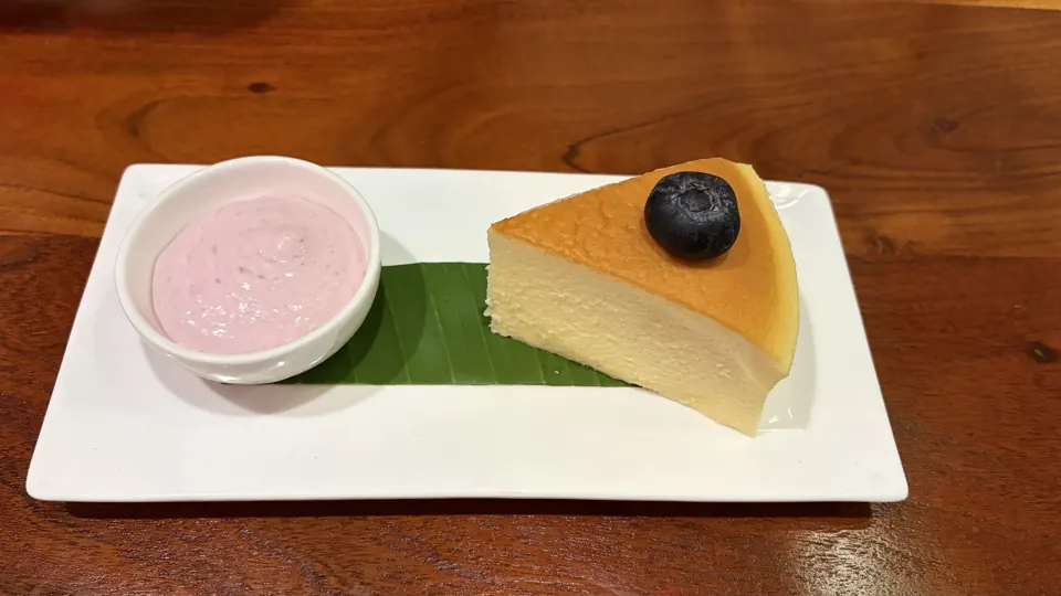 Slice of fluffy cheesecake with a blueberry on banana leaf beside a bowl of pink cream